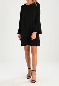 Svart, långärmad klänning med rund hals, avslappnad passform, knälång nederdel och utsvängda ärmar. Kombineras med svarta sandaletter med remmar.