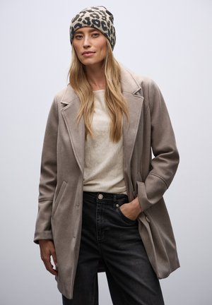 Manteau en mélange de laine gris clair avec des revers, deux poches, porté sur un pull crème et un jean noir. Accentué par un bonnet léopard gris et noir.