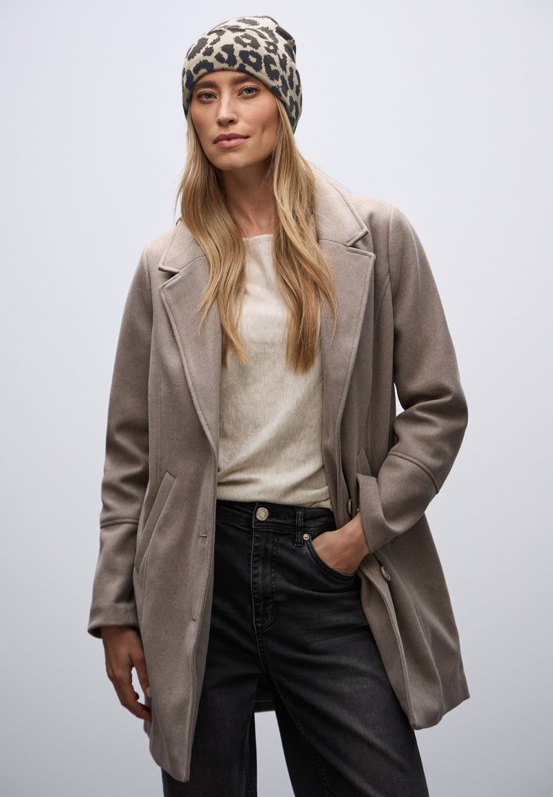 Cappotto in misto lana grigio chiaro con revers, due tasche, indossato sopra un maglione crema e jeans neri. Accentuato con un berretto leopardato grigio e nero.