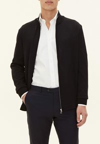 Black zip-up jacket with ribbed collar and cuffs, made of smooth fabric, paired with a white shirt and dark trousers, hands in pockets.