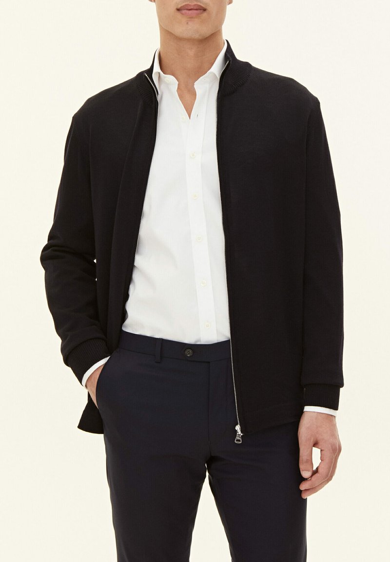 Black zip-up jacket with ribbed collar and cuffs, made of smooth fabric, paired with a white shirt and dark trousers, hands in pockets.