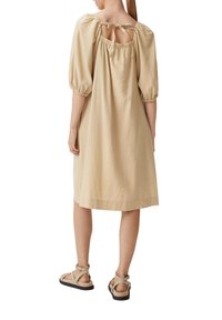 Vestito in lino beige con vestibilità rilassata, maniche a sbuffo e dettaglio con laccetto sul retro. Indossato con sandali piatti e a strappo. Texture liscia e lunghezza fino al ginocchio.