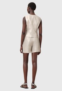 Femme portant un haut beige sans manches et un short assorti avec des sandales argentées, debout de dos sur un fond clair uni.
