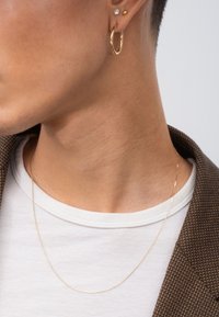 Goldene Creolen und eine minimalistische goldene Kettenhalskette an einem Modell. Der Hautton ist hell mit einer glatten Textur vor einem weißen Hintergrund.