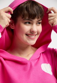Maglione rosa con cappuccio, caratterizzato da un design a cuore bianco in rilievo sul petto. Il tessuto sembra morbido con una consistenza liscia.
