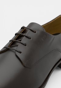 Scarpa in pelle marrone scuro con finitura liscia, design senza cuciture e sei occhielli per i lacci con dettagli di cucitura a contrasto.