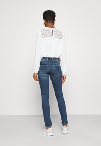 Vit topp med spetsdetaljer på baksidan, kombinerad med blå smala jeans och vita sneakers. Outfiten har en avslappnad design.