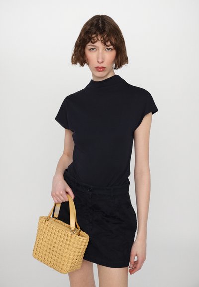 Svart kortermet topp med høy hals, parret med mørke shorts. Holder en liten, vevd gul håndveske med beige hanker.