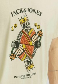 Kreminė marškinėlių su spalvinga grafika, kurioje pavaizduota karūnuota skeletas, laikančiais gėlę, su dekoratyviniais raštais ir tekstu "JACK & JONES 1990".