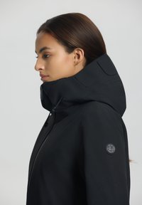 Veste à capuche noire avec un col haut, fermeture éclair et un écusson rond sur la manche. Texture lisse et design moderne.
