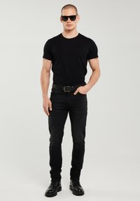 Bărbat în picioare, cu fața înainte, purtând ochelari de soare negri, tricou negru, blugi negri, centură neagră și cizme negre pe un fundal simplu.