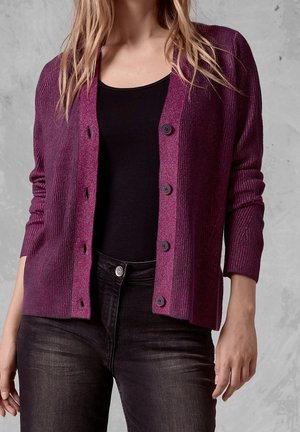 Woman wearing a ribbed purple buttoned cardigan over a black top and black jeans, standing against a gray textured background.