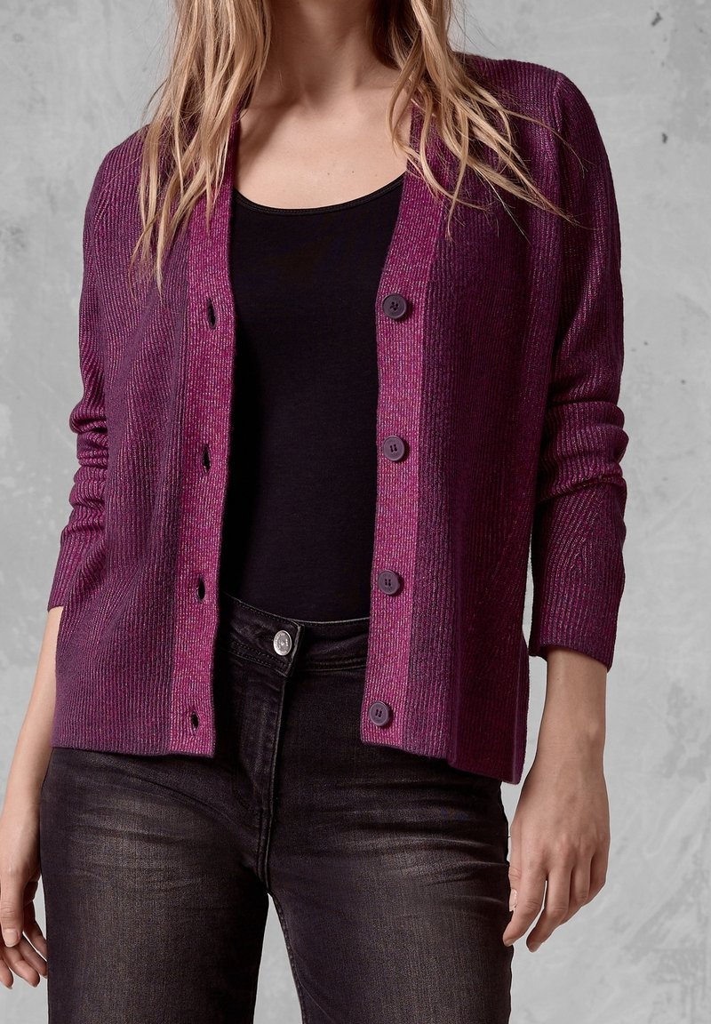 Femme portant un cardigan violet côtelé à boutons par-dessus un haut noir et un jean noir, debout devant un fond texturé gris.