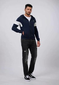 Sudadera con cremallera en color navy con rayas blancas, combinada con jeans oscuros desgastados y zapatillas negras. Ajuste estructurado, tejido suave y diseño casual.