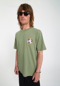 Homme portant des lunettes de soleil noires, un t-shirt vert avec un emblème de globe, un pantalon noir et un bracelet tressé, debout devant un fond uni.