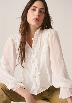 Tânără cu păr lung și castaniu, purtând o bluză albă cu volane și pantaloni kaki cu broderie florală, așezată pe un fundal simplu.