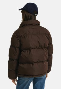 Bruine pufferjack met een hoge kraag en quilted details, gecombineerd met een marineblauwe pet. Zichtbare stiksels en een plush textuur verhogen de warmte.