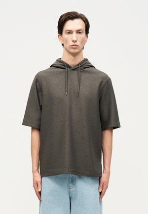 Mann trägt eine dunkelgraue Kapuzenjacke mit kurzem Ärmel und dezentem Muster sowie helle, weit geschnittene Jeans, steht vor einem einfarbigen weißen Hintergrund.