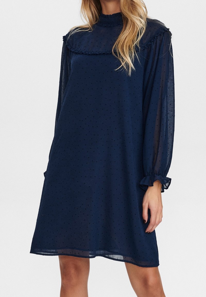 Femme portant une robe bleue marine transparente longueur genou avec manches longues, col haut à volants et motif à pois subtil.