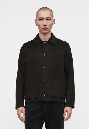 Homme au crâne rasé portant une veste noire boutonnée avec trois poches avant et un pantalon noir sur un fond uni.