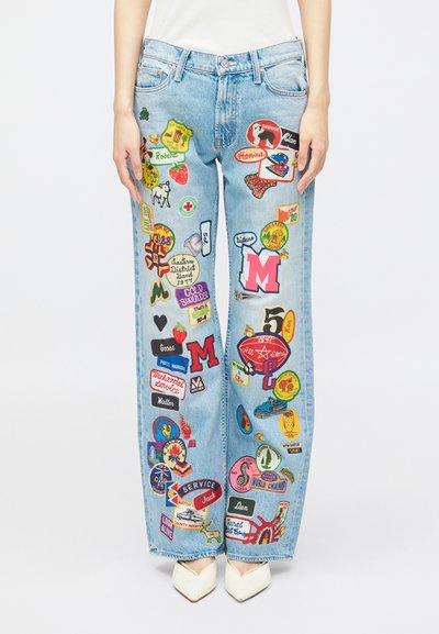 Jeans azzurri chiari con diverse toppe ricamate colorate, abbinati a scarpe bianche con punta e a una maglietta bianca, su uno sfondo bianco.