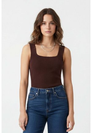 Jeune femme aux cheveux bruns ondulés portant un haut marron sans manches en maille côtelée et un jean bleu taille haute, sur un fond blanc uni.