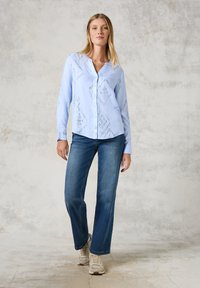 Hellblaues Button-up-Hemd mit geometrischen Ausschnittmustern, langen Ärmeln und Kragen. Kombiniert mit blauen Jeans und beigen Sneakern.