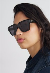 Occhiali da sole oversized neri con montatura geometrica, lenti scure e un accento dorato sul lato, su uno sfondo neutro.