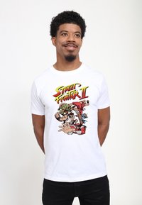 Bílý bavlněný tričko s barevným grafikou "Street Fighter II", zobrazující postavy v dynamických pózách, s tučným nápisem nahoře.