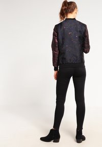 Svart bomberjacka med mönstrad tyg, med glänsande ärmar och ribbad nederkant, kombinerad med svarta slim fit jeans och ankelboots.