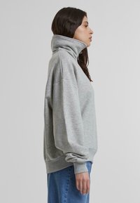 Sudadera gris claro con cuello alto, hombros caídos y puños acanalados, combinada con jeans azul. Tejido suave y texturizado.