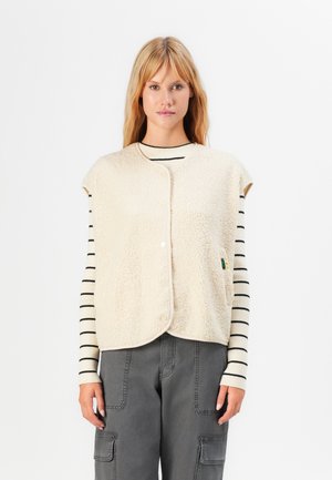 Femme portant un gilet à boutons-pression en sherpa crème sur un haut à rayures crème et noir à manches longues et un pantalon cargo gris, se tenant devant un fond blanc.