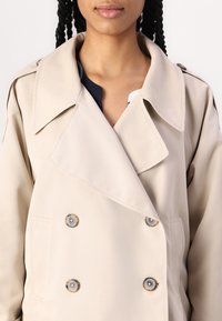 Vaaleanbeige kaksirivinen trench coat, jossa on suuret kaulukset, metallinappiset kiinnitykset ja sivutaskut. Sujuva kangas, jossa on hieman kiiltävä pinta.