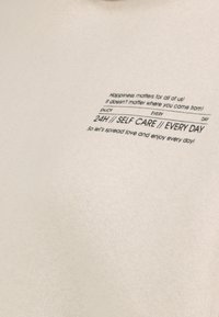 Beige katoenstof met een zachte textuur, voorzien van bedrukte tekst: "Geluk is belangrijk voor ons allemaal!" en "24u // ZORG VOOR JEZELF // ELKE DAG."