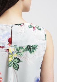 Bloemenloze mouwloze blouse van lichtblauwe stof. Beschikt over een keyhole rug met knoopsluiting en meerkleurige bloemenpatronen.