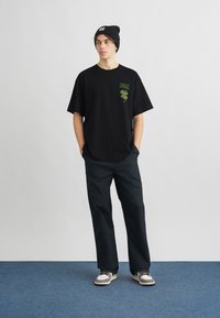 Czarna koszulka z zielonym graficznym wzorem koniczyny i napisem, zestawiona z ciemnymi spodniami i trampkami. Model nosi czarną czapkę beanie.