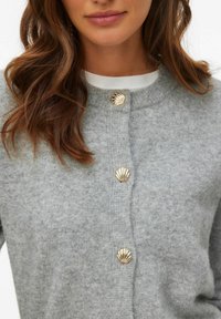 Grå stickad kofta med dekorativa gyllene knappar i form av snäckskal, rund halsringning och medeltexturerat tyg.