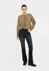 Dugi top dugih rukava s leopardovim uzorkom u bež i smeđoj boji, uparan s crnim trapez hlačama i crnim cipelama. Model stoji s neutralnim izrazom lica.