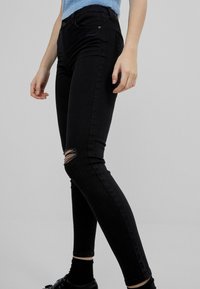 Jeans skinny negros con detalle de rodilla desgastado y una textura suave. Cintura de altura media con cierre de botón y diseño clásico de cinco bolsillos.