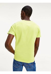Camiseta amarilla brillante de algodón, con cuello redondo y mangas cortas. Lleva puesta con pantalones vaqueros azules, vista desde la parte de atrás.