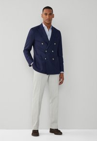 Marinblå dubbelknäppt kavaj med guldknappar, parat med en ljusblå skjorta och skräddarsydda krämfärgade byxor, avslutat med bruna loafers.