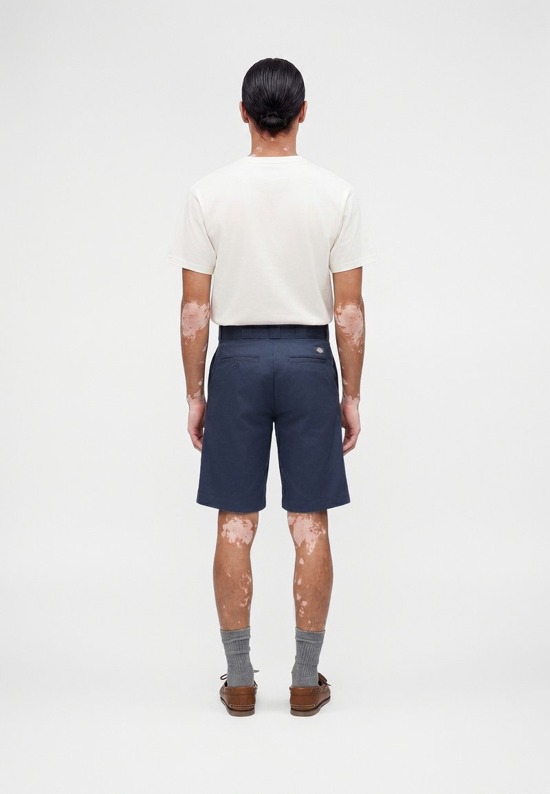 Mann mit Vitiligo steht mit dem Rücken zur Kamera, trägt ein weißes kurzärmeliges Hemd, dunkelblaue Shorts, graue Socken und braune Schuhe vor einem einfarbigen Hintergrund.