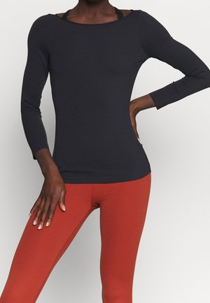 Vrouw draagt een nauwsluitende zwarte top met lange mouwen en gebrande oranje leggings, met hand rustend op de heup tegen een effen lichte achtergrond.