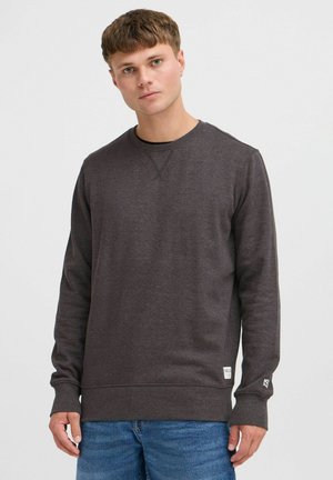Sudadera gris oscuro de manga larga, cuello redondo, con puños y dobladillo acanalados. Cuenta con un logo en la manga y una pequeña etiqueta en la parte inferior izquierda.