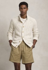 Cardigã creme com malha de cabos e botões de madeira, combinado com calções caqui. Mistura de algodão, corte descontraído, acabamento texturizado e punhos dobrados.
