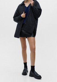 Femme portant une grande veste noire, un pull bleu marine, une mini-jupe en cuir noir, des chaussettes noires et des mocassins épais noirs sur fond blanc.