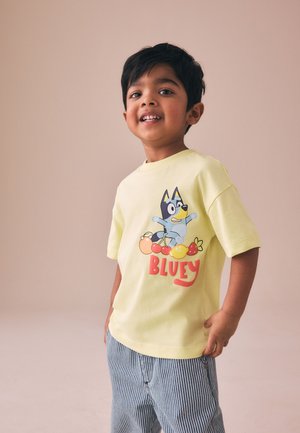 Smilende ung dreng iført en gul t-shirt med frugtillustrationer og stribede bukser, stående mod en ensfarvet baggrund.
