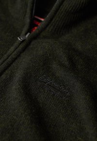 Maglione zip con zip di colore verde scuro realizzato in un tessuto texturizzato. Presenta un logo ricamato nero ben visibile sul davanti e un colletto a coste.