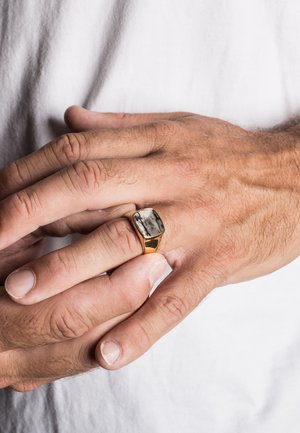 Gold ring with a rectangular gray and black stone on a hand with visible texture and skin tone variations, positioned against a white fabric.