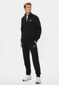 Felpa nera con zip e pantaloni abbinati in morbido tessuto, con il logo EA7 bianco sul petto e sulla coscia. Scarpe da ginnastica bianche.